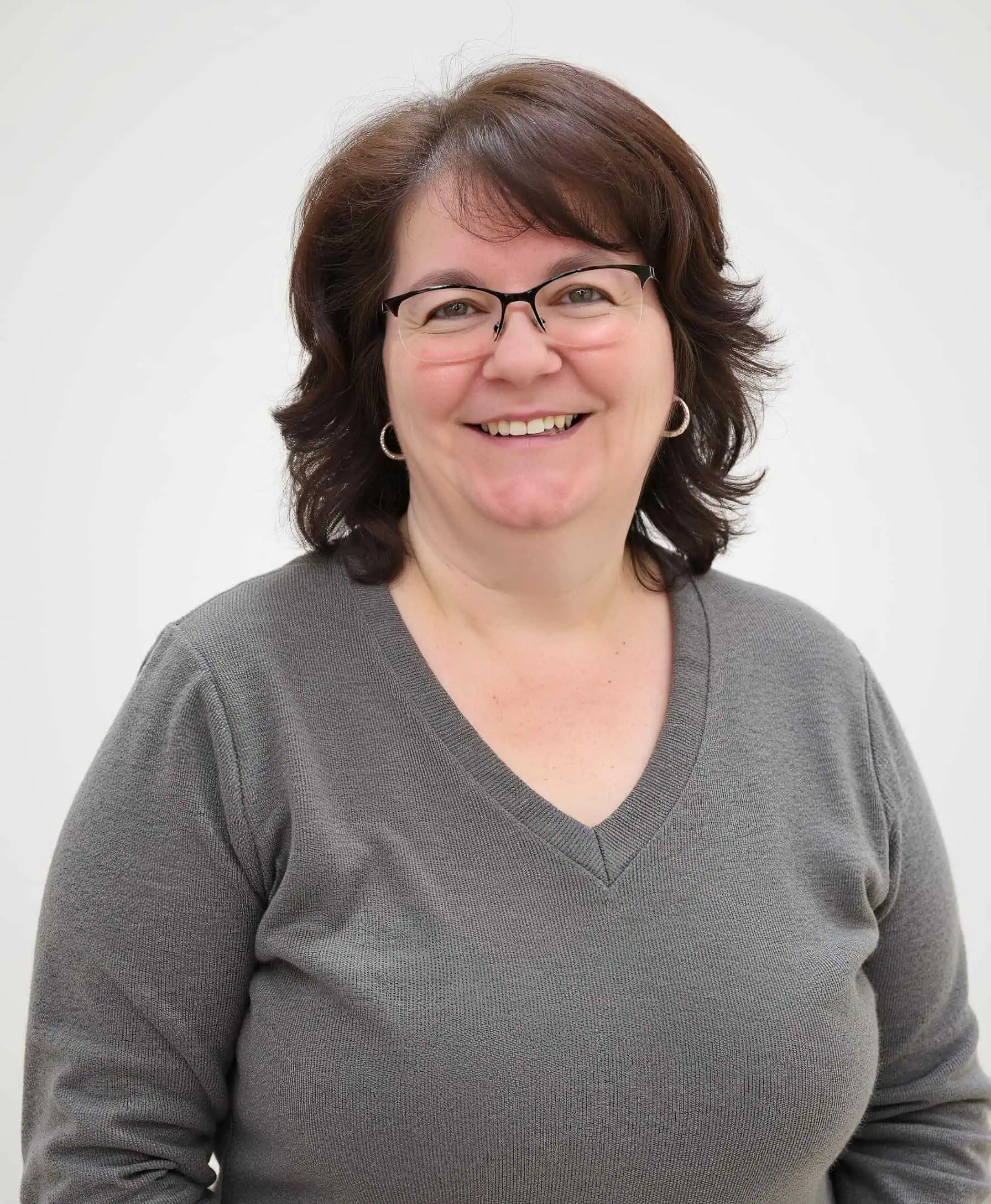 Smiling woman in a gray V-neck sweater with shoulder-length brown hair and glasses, standing in front of a bright white background.